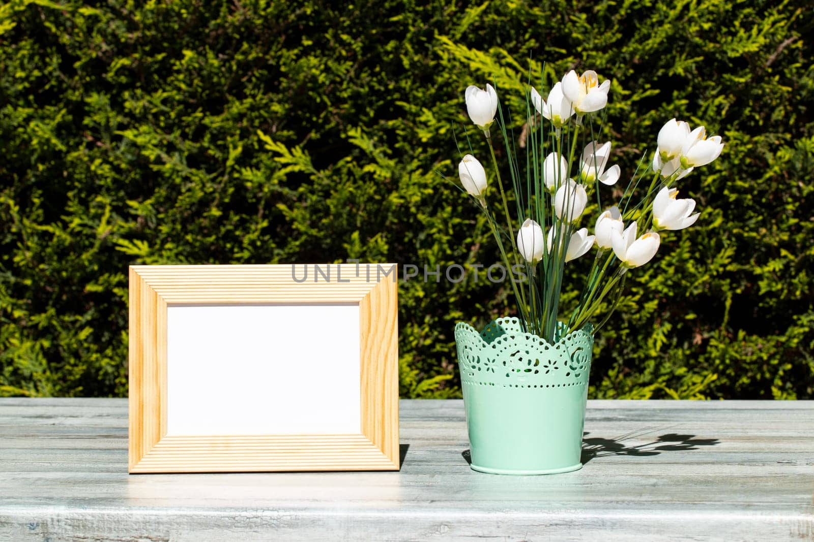Empty wooden frame and white wild flowers on rustic bright wooden table over natural green background. Space.