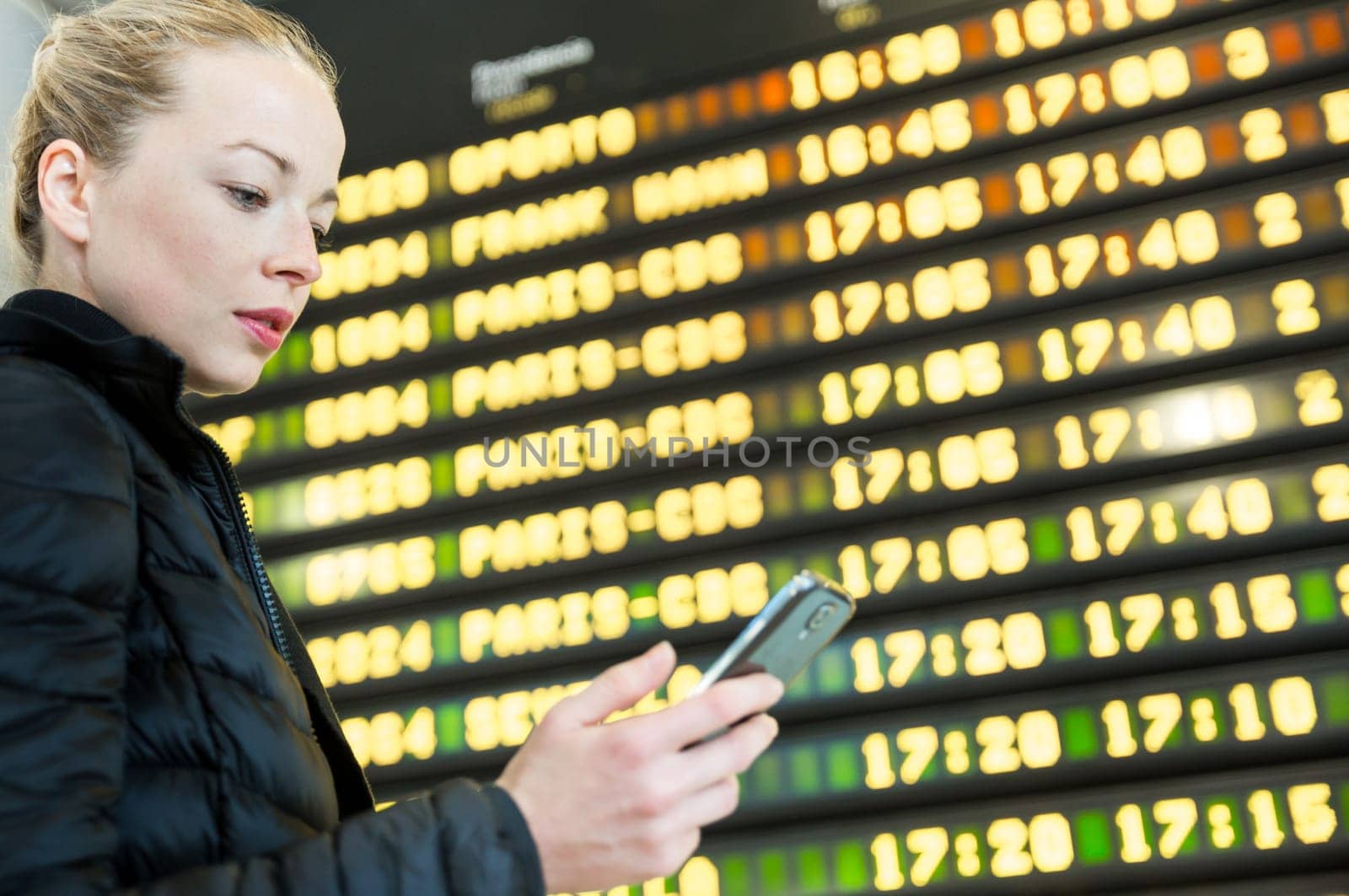 Young woman at international airport looking at the flight information board, holding smart phone in her hand, checking flight informations on phone application.