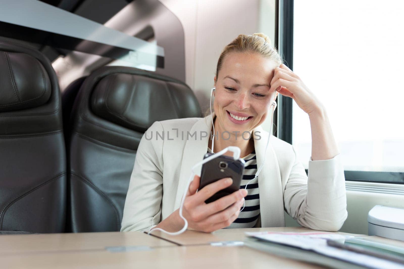 Businesswoman communicating on cellphone using headphone set while traveling by train in business class seat.