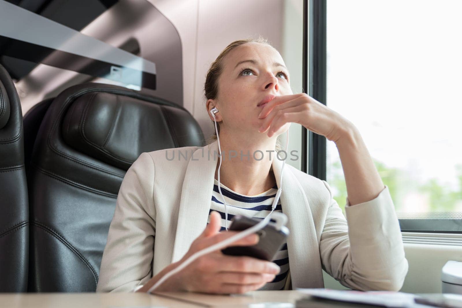 Thoughtful businesswoman communicating on cellphone using headphone set while traveling by train in business class seat.