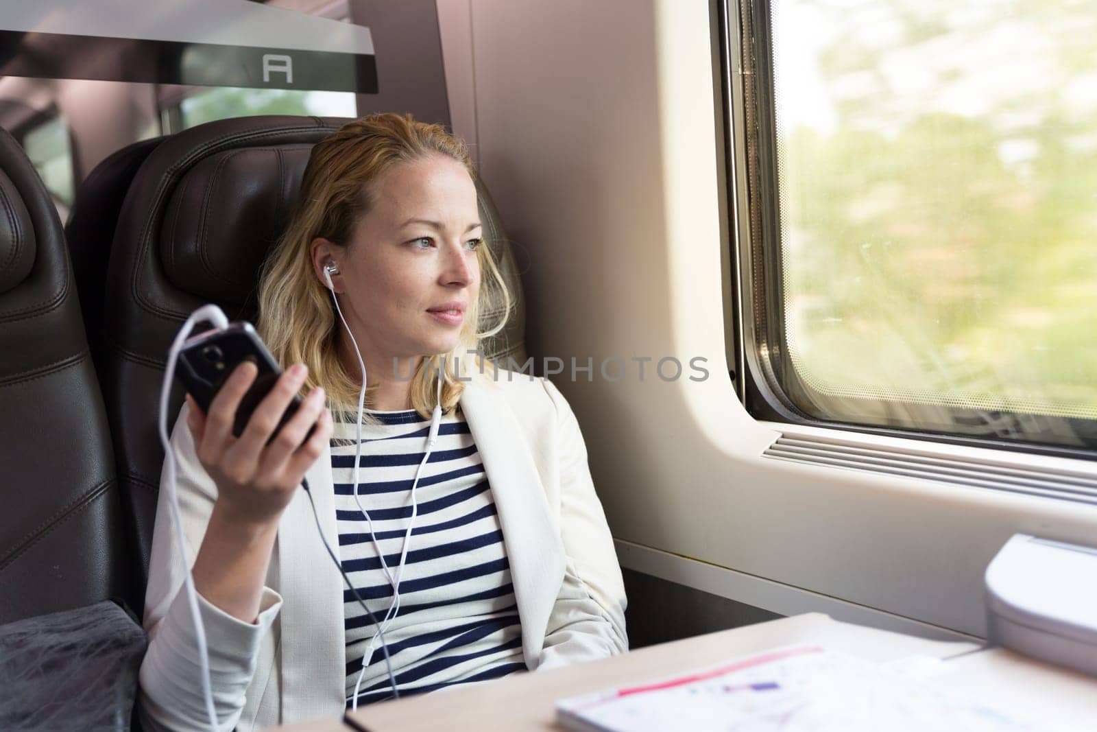 Businesswoman communicating on cellphone using headphone set while traveling by train in business class seat.