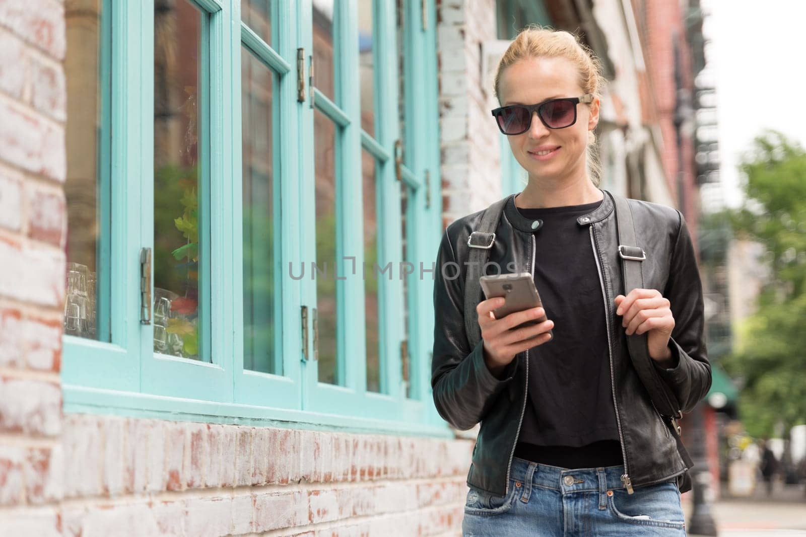 Fashionable Young Woman in Black Leather Jacket and Busy with her Mobile Phone While Walking a City Street of New York City.
