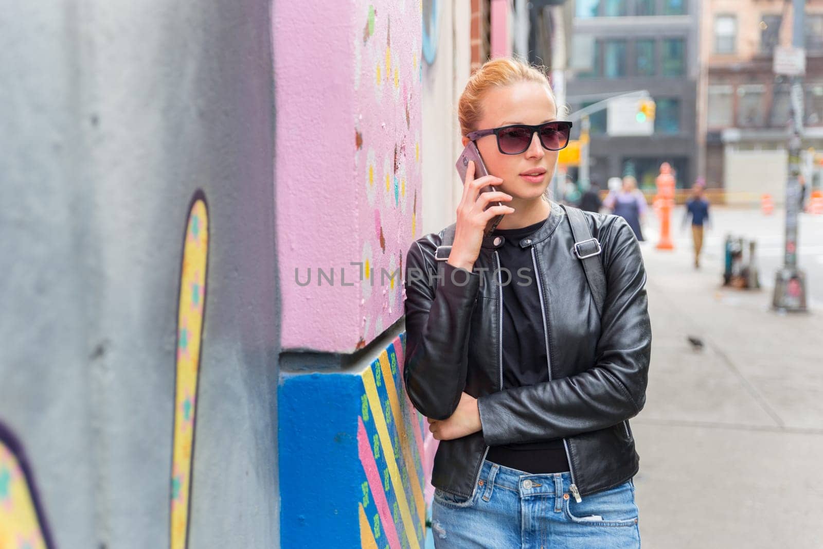 Stylish woman talking on smartphone leaning against colorful graffiti wall in East Village, New York city, USA.