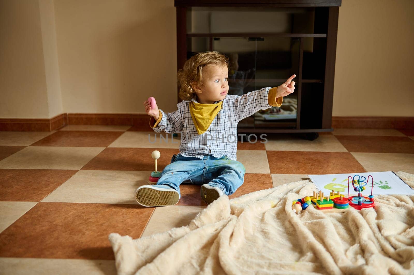 A toddler sits on the floor indoors surrounded by toys, expressing curiosity and joy while pointing towards something with a focused look.