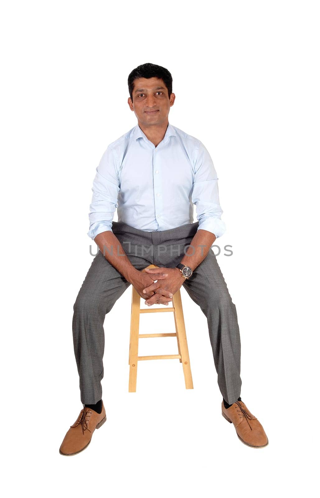 A good looking East Indian man in a suit pants and light blue shirt.sitting on a bar chair, isolated for white background.