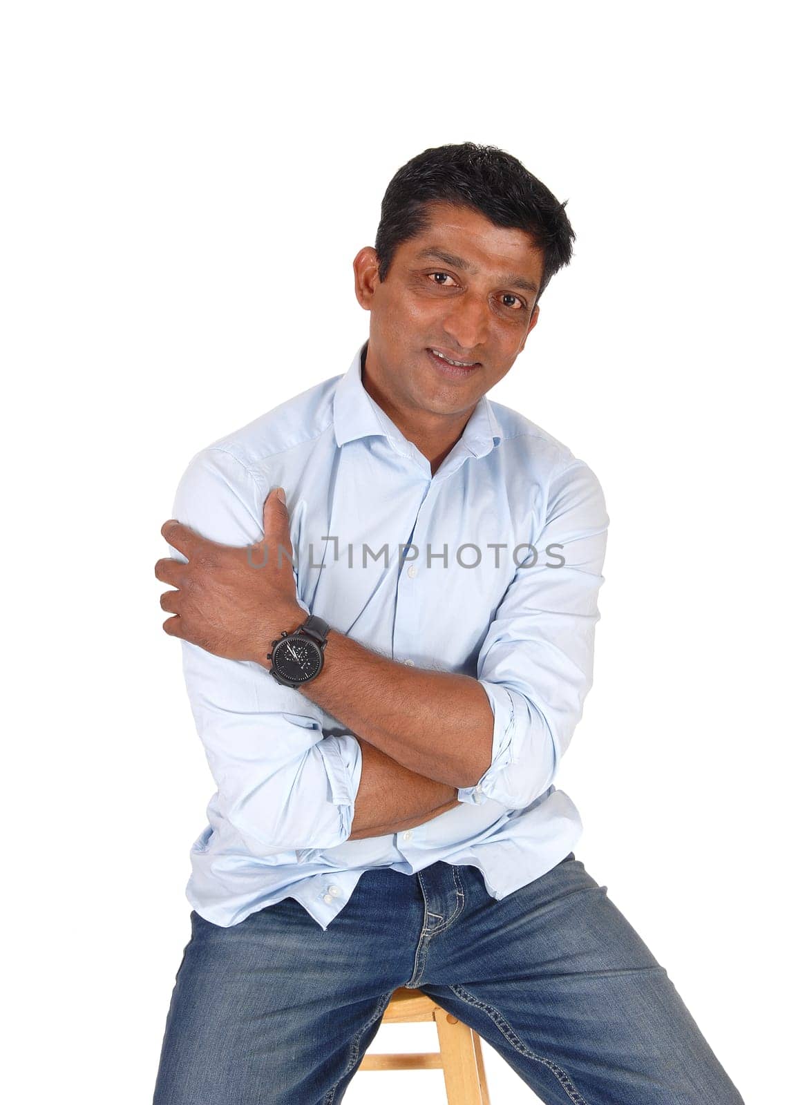 A handsome middle age East Indian man sitting on a bar chair in.jeans and a blue shirt, isolated for white background.