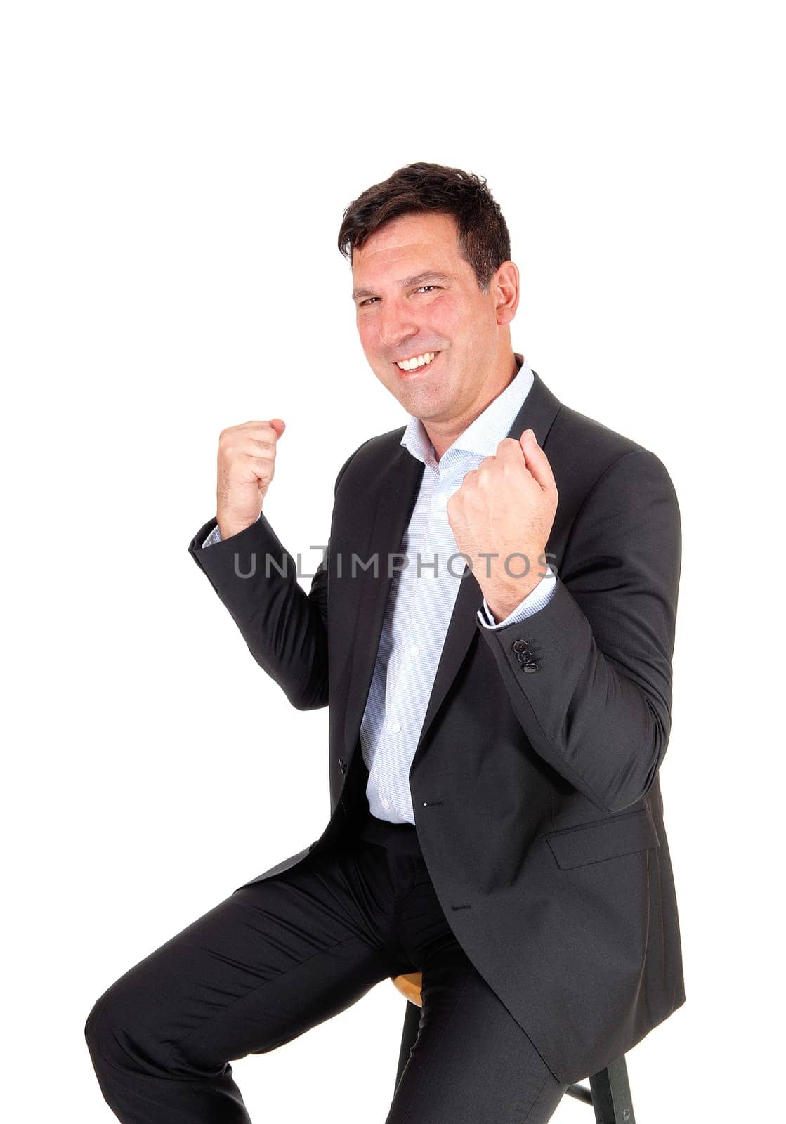 A happy handsome middle age man sitting in a suit with his hands to .fists, smiling over his victory, isolated for white background.