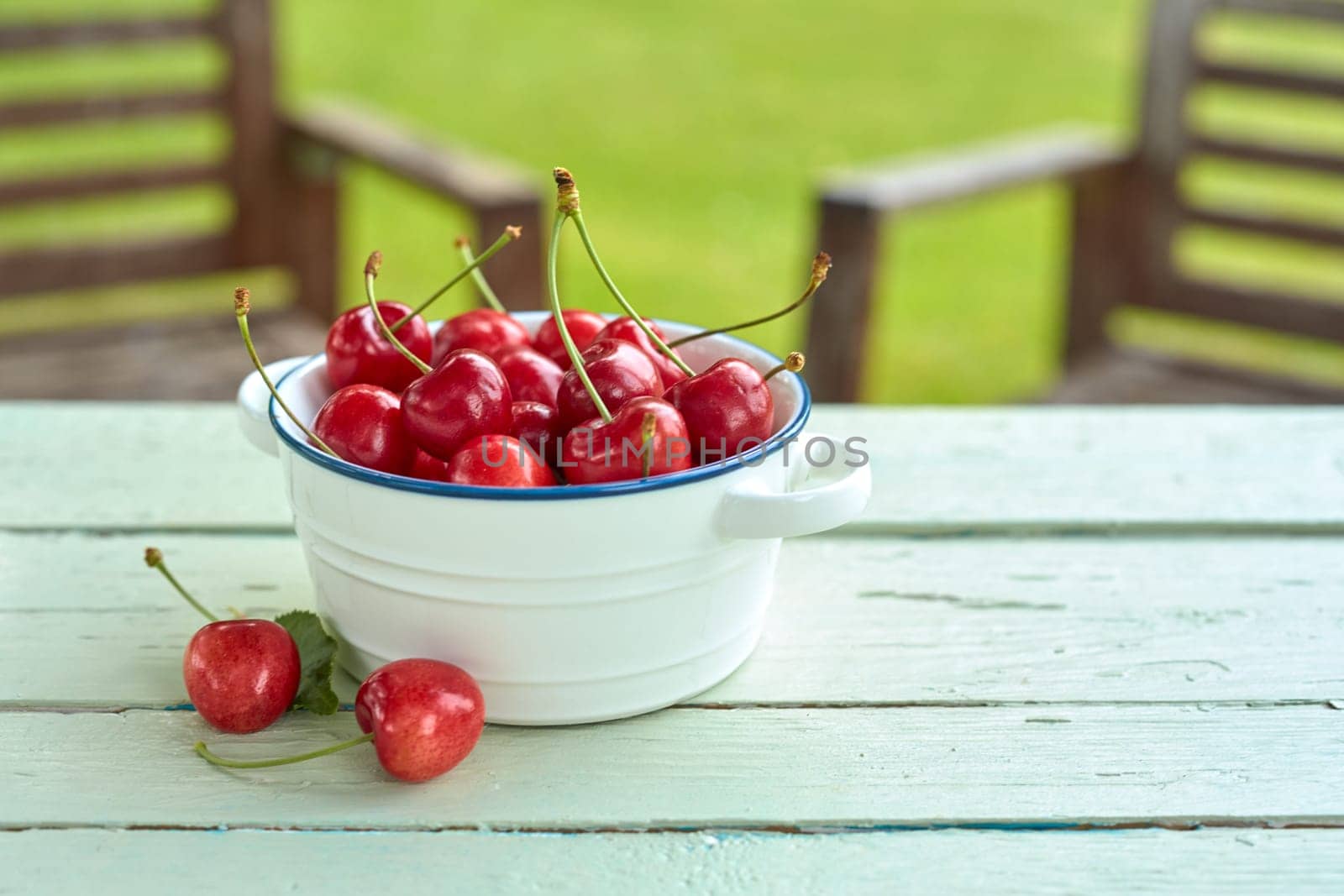 Dish of freshly harvested red sweet cherries on a rustic white garden table with green lawn and chairs in summer