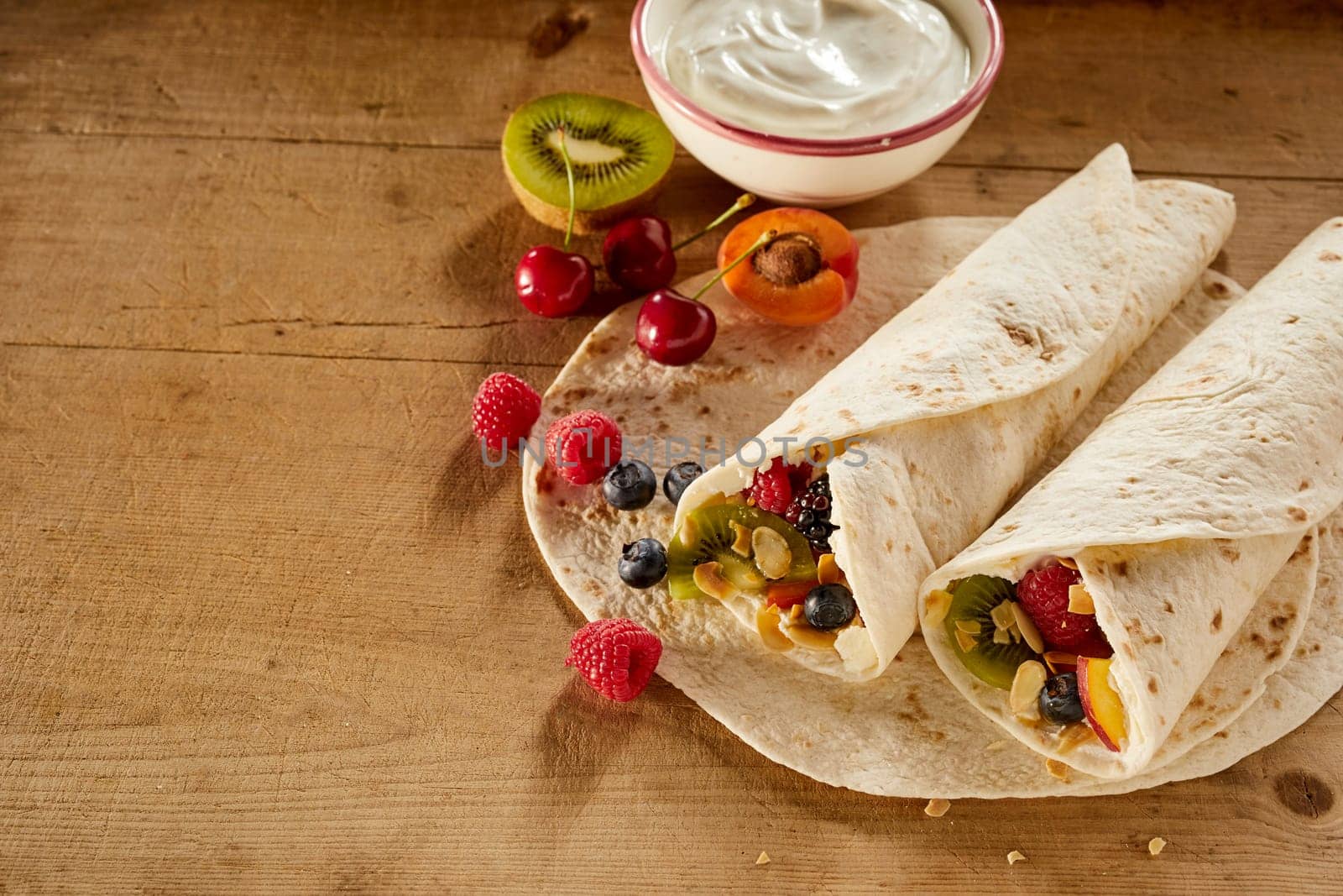 Assorted fresh tropical fruit in a wrap or pancake with whipped cream including kiwi, raspberry, blueberry, apple, apricot and cherries viewed high angle on a wooden table with copy space