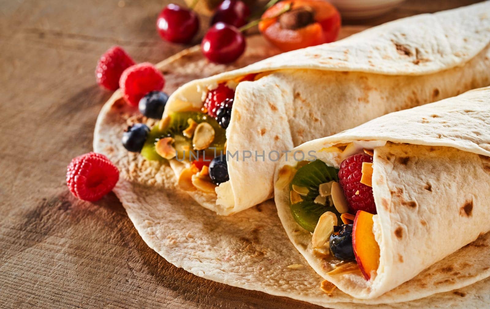 Close up on two healthy fresh fruit and nut wraps with assorted tropical summer fruit and whipped cream on a wooden table with scattered ingredients