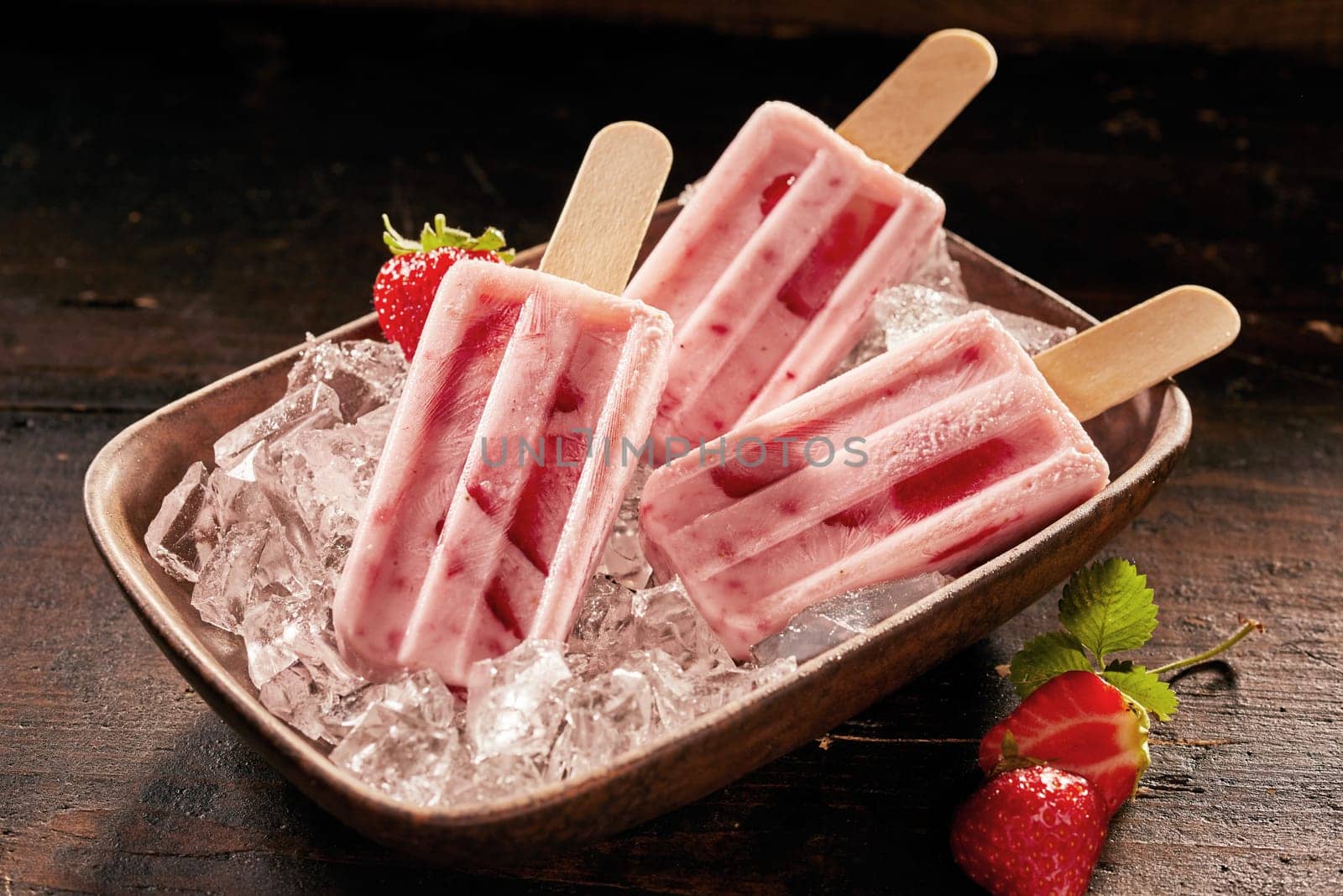 Dish of healthy fresh strawberry popsicles or frozen suckers on ice with scattered berries on a rustic wooden table for a refreshing summer treat