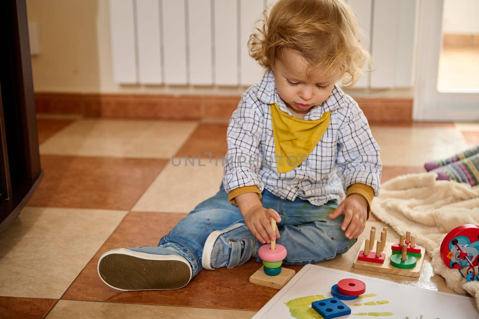 A young child sitting on the floor next to educational toys, demonstrating cognitive and motor skills development inside a bright living room.