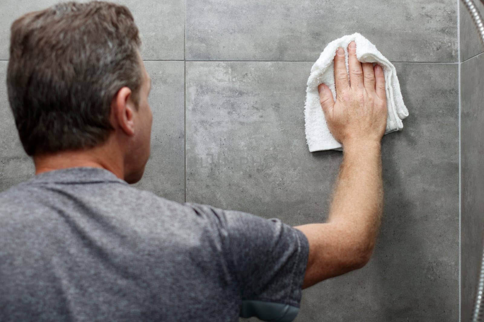 Male scrubbing a home bathroom gray tile shower wall with his hand holding a wet white cotton rag. White cotton cloth being used by an older caucasian man to clean a bathroom shower gray tile wall.