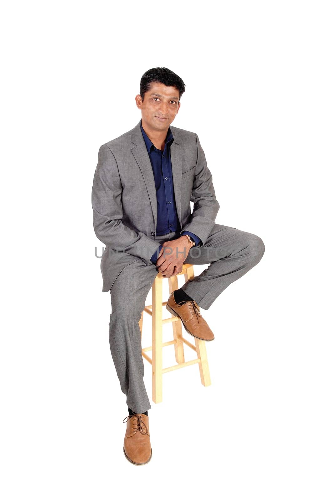 A middle age East Indian business man sitting in a gray suit and blue.shirt on a bar chair, isolated for white background.