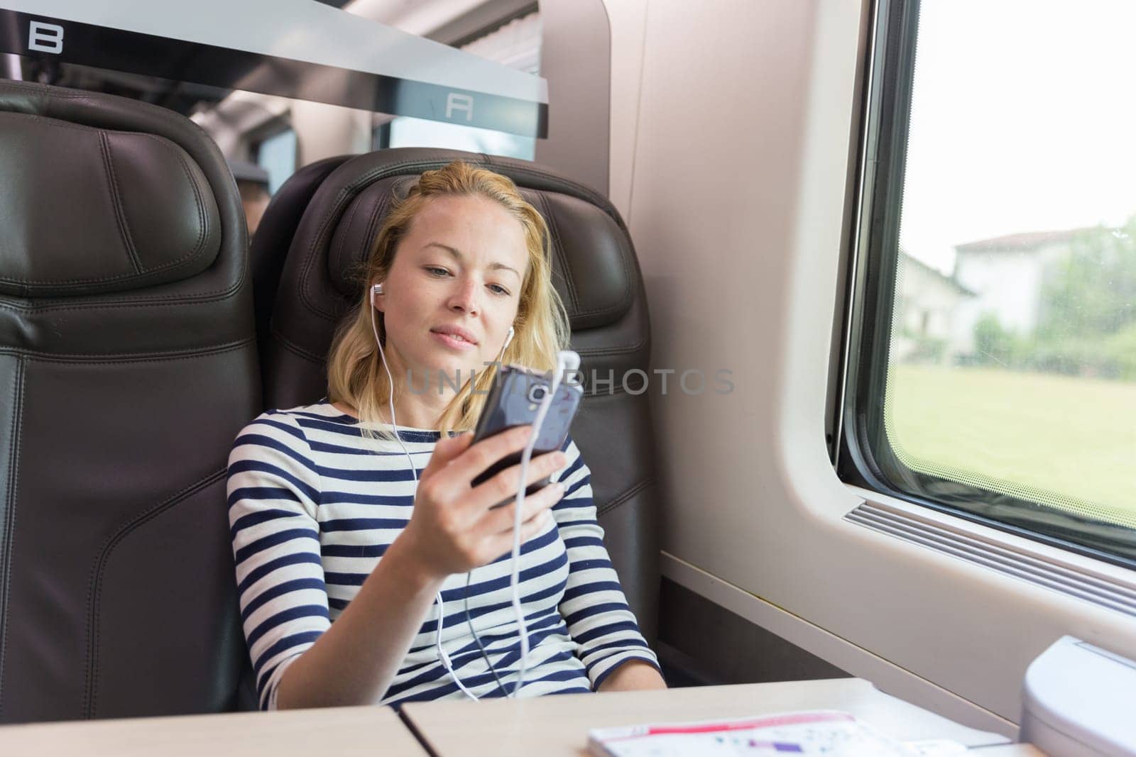 Businesswoman communicating on cellphone using headphone set while traveling by train in business class seat.