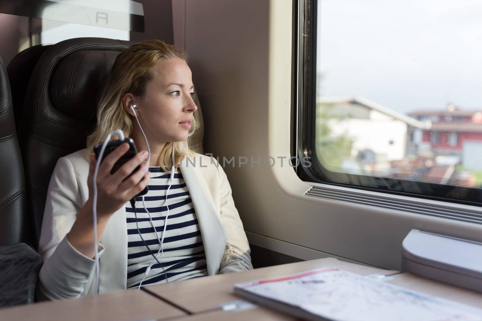 Thoughtful businesswoman looking trough the window, listening to podcast on cellphone using headphone set while traveling by train in business class seat.