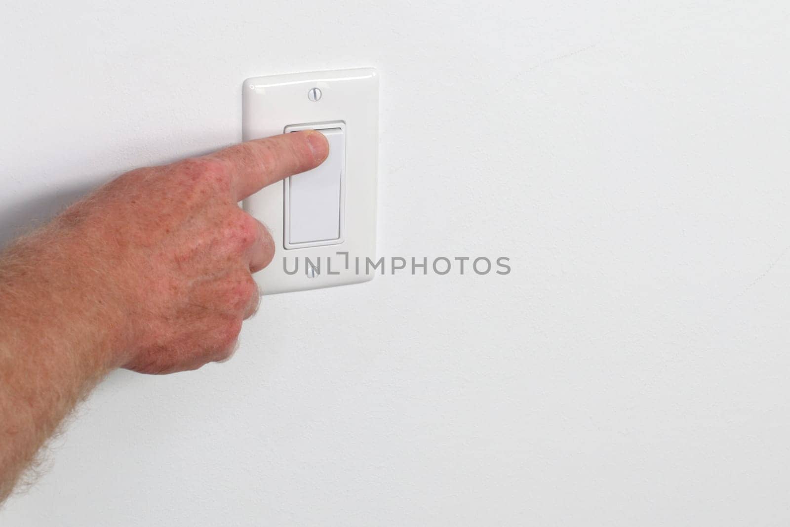 One adult male hand pressing a light switch panel. One flat white panel light switch being turned on Male hand pressing on a flat white wall switch panel from the left to turn on the light in a room.