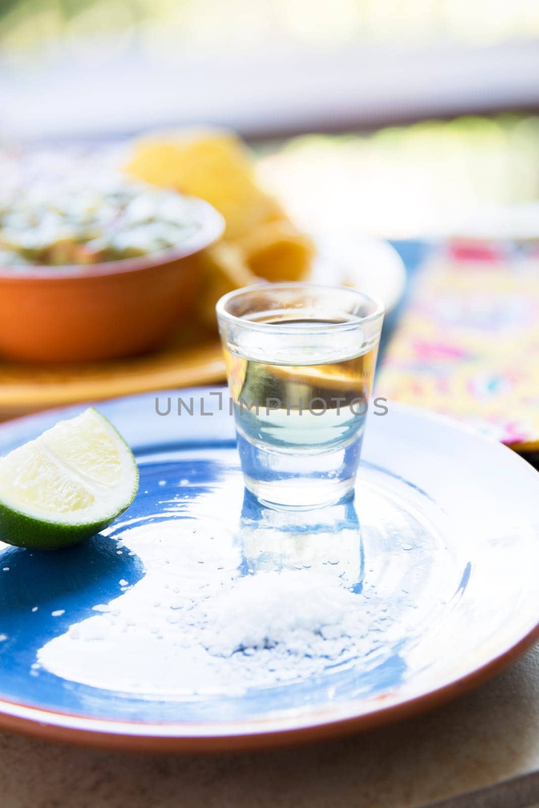 Shot of tequila on blue plate with lime and salt, chips and guacamole out of focus in the background