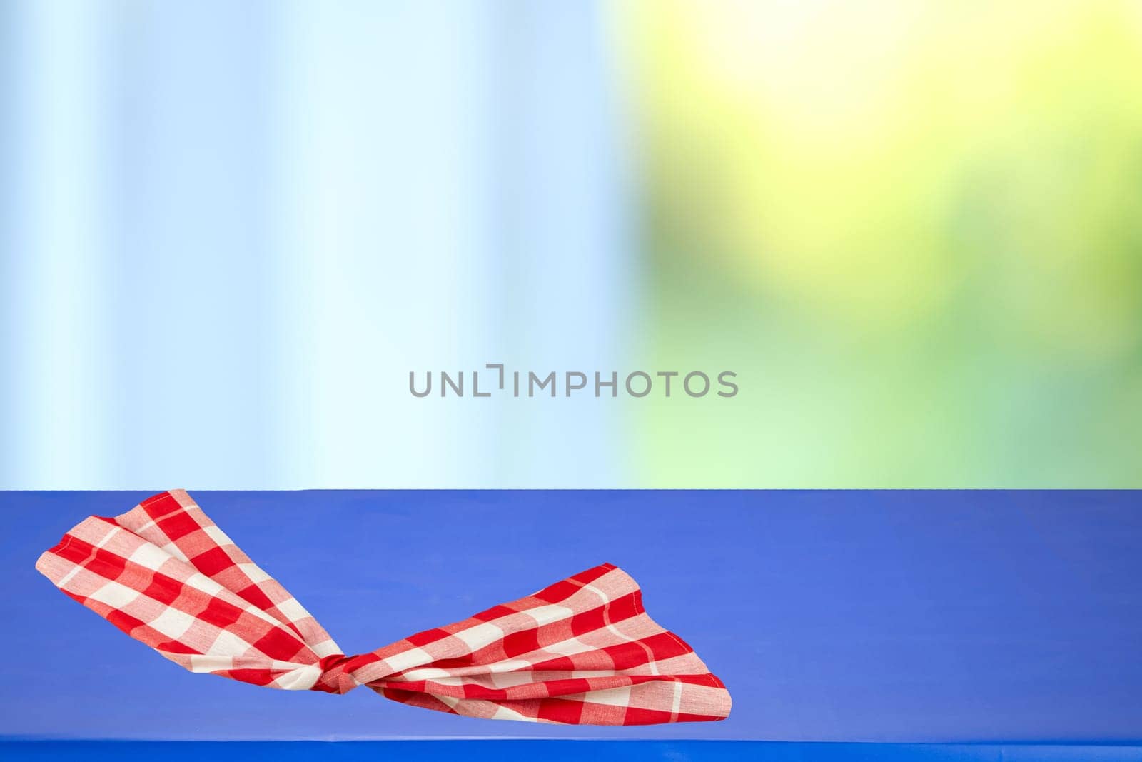 Red checkered tablecloth folded on a blue table with blurred bright pastel background. Space for your food and product display montage.