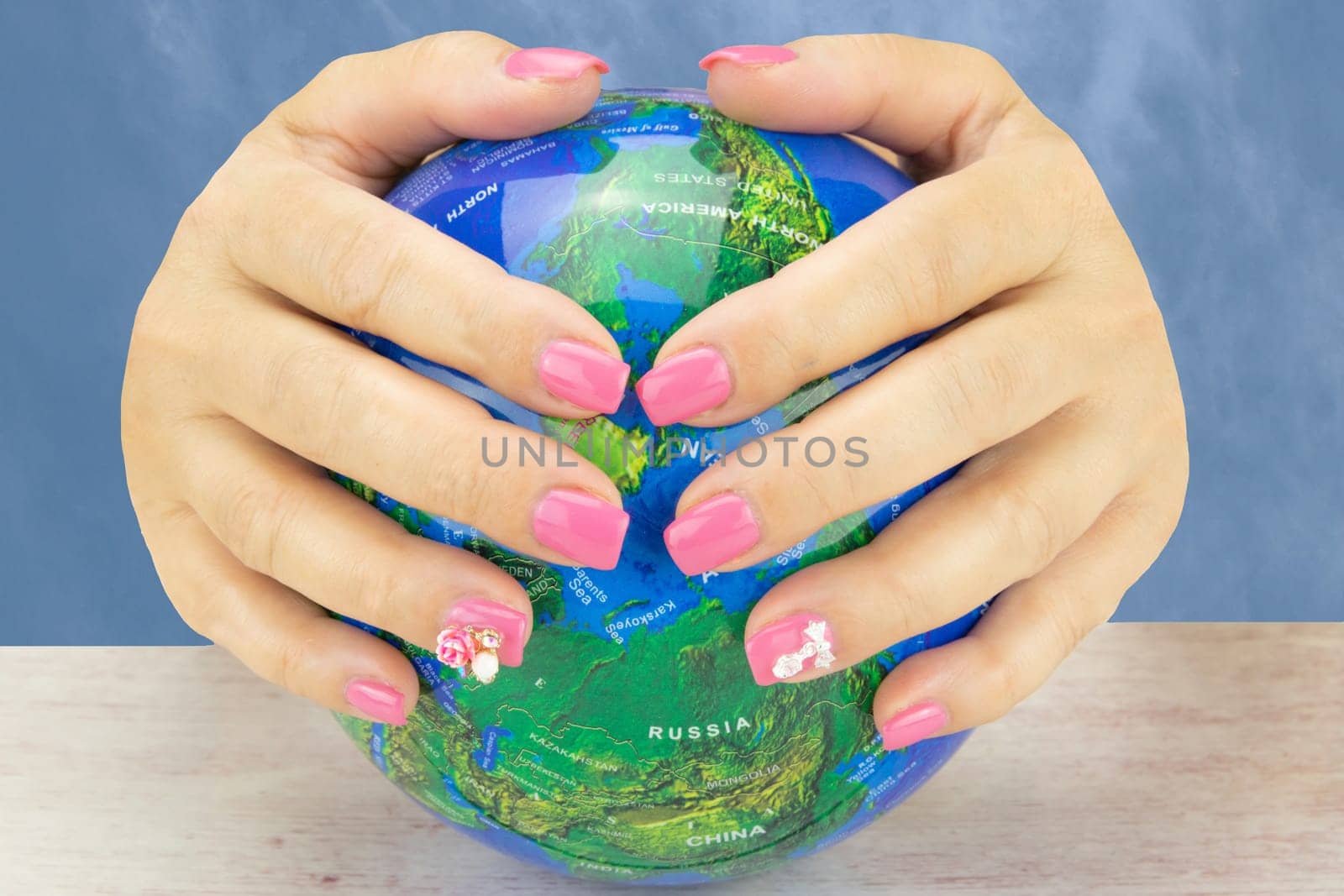 Closeup of beautiful pink nail design at female hands at globe on a wooden table. One figure on one nail at each hand decorate the pink nail design. Background is a natural blue sky. Fashion nail design manicure.