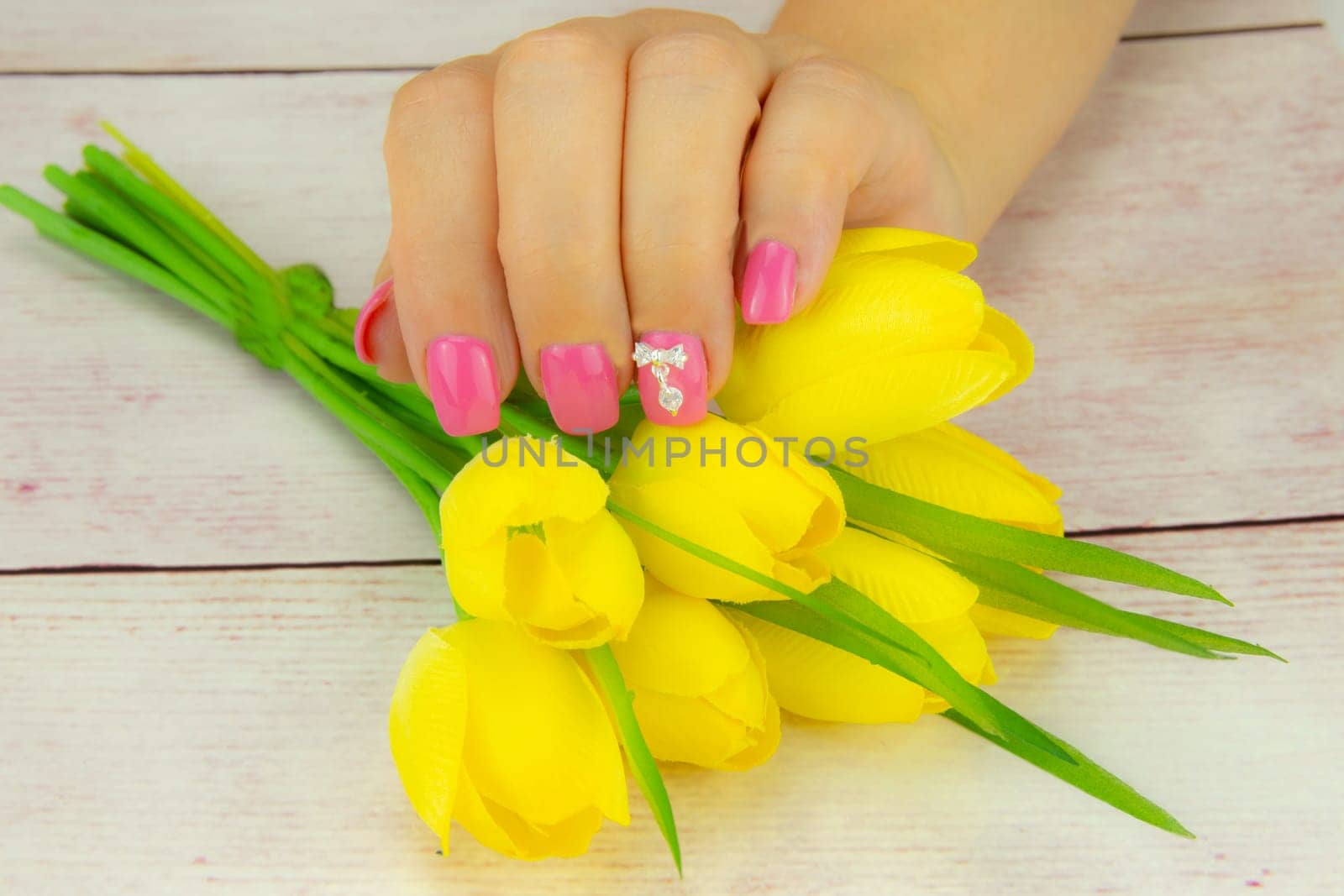 Female hand of woman, pink manicured on nails with a bouquet of yellow tulips. A figure on a nail decorate the pink nail design. Background is a rustic bright table. Concept beauty.