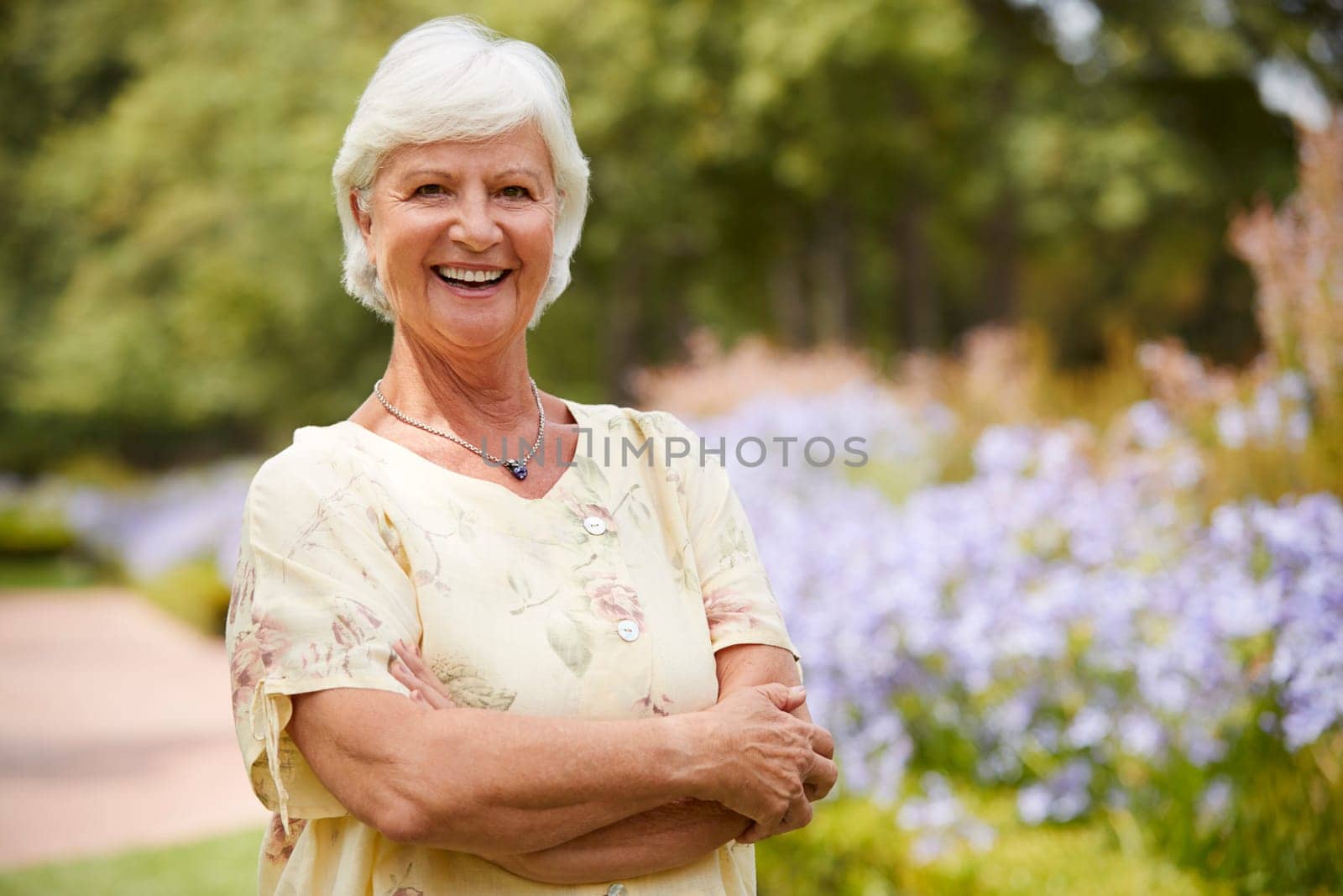 Portrait Of Senior Woman Enjoying Walk By Flower Beds In Park