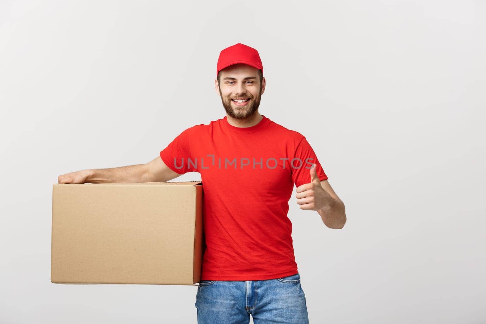 Cheerful young handsome delivery man holding a cardboard box and showing his thumb up while standing against grey background.