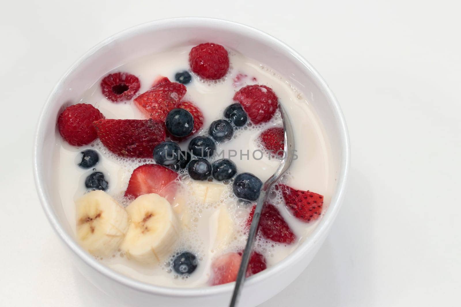 White breakfast bowl of banana, strawberries, blueberries, raspberries, almond milk and cereal. Morning meal of mixed berries, banana, almond milk and organic cereal in a white dish with a spoon..