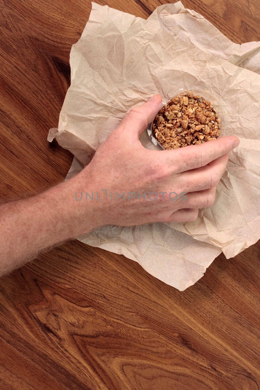 Granola mix treat in a small clear plastic container being held by an adult male hand. Sweet granola snack mixture in a tiny plastic bowl held by fingers of a male hand over a light brown napkin.