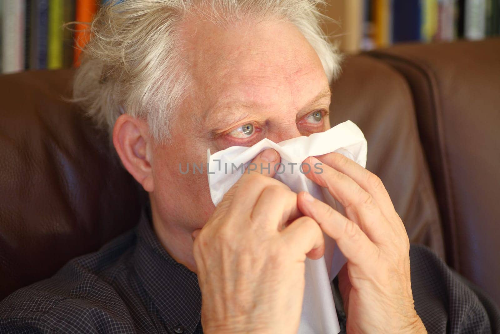 Older man blows his nose with a tissue while seated on a sofa