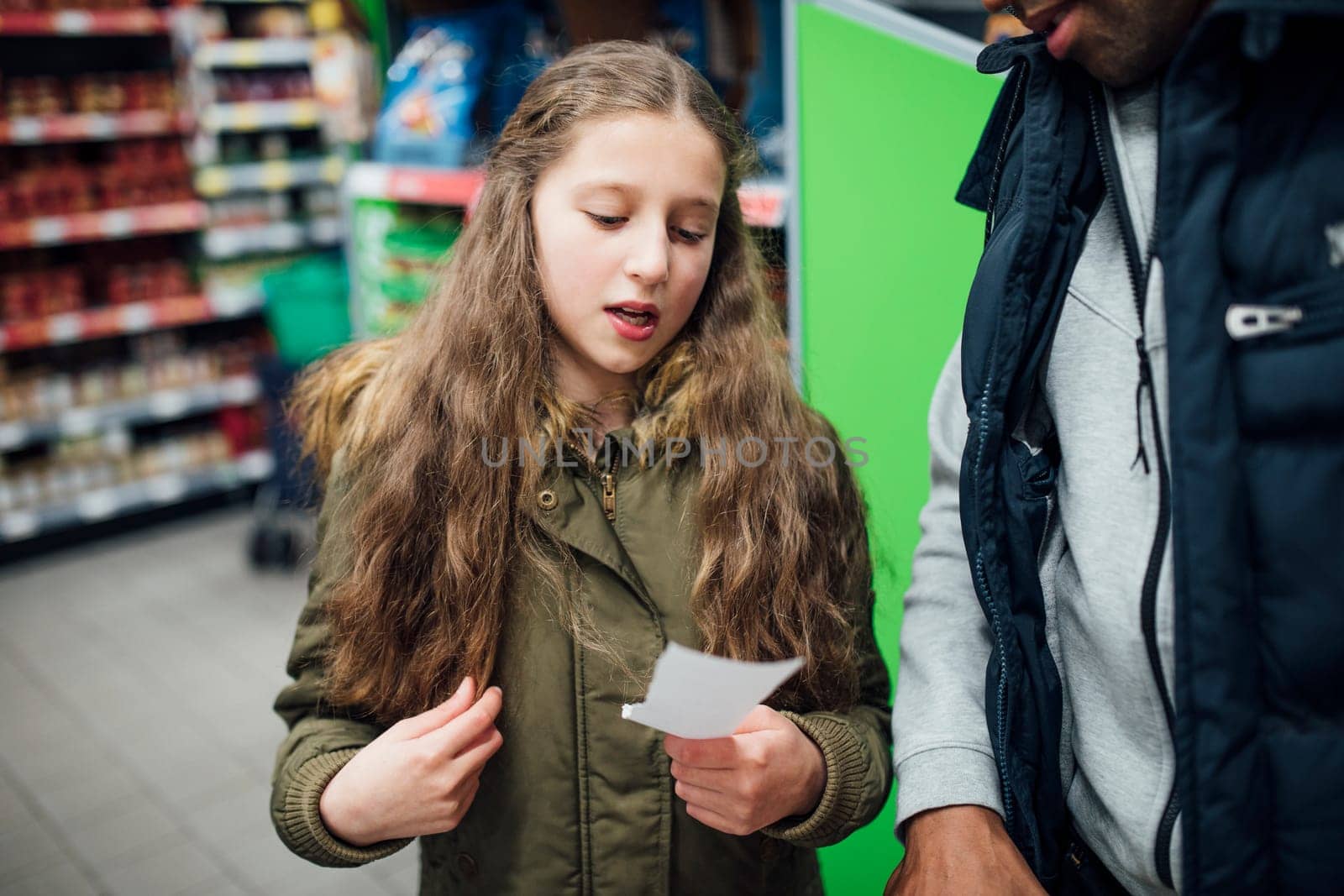 Little girl is reading things from a shopping list to her father in the supermarket.