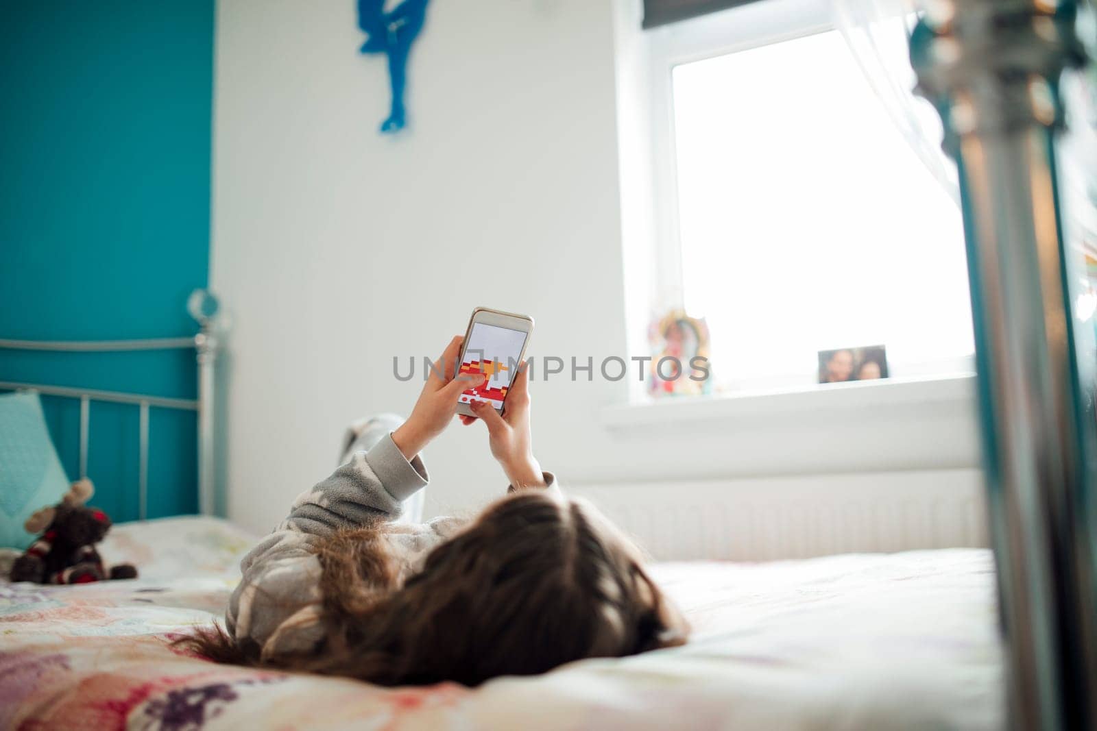 Little girl is lying on her bed, playing a game on her smartphone.