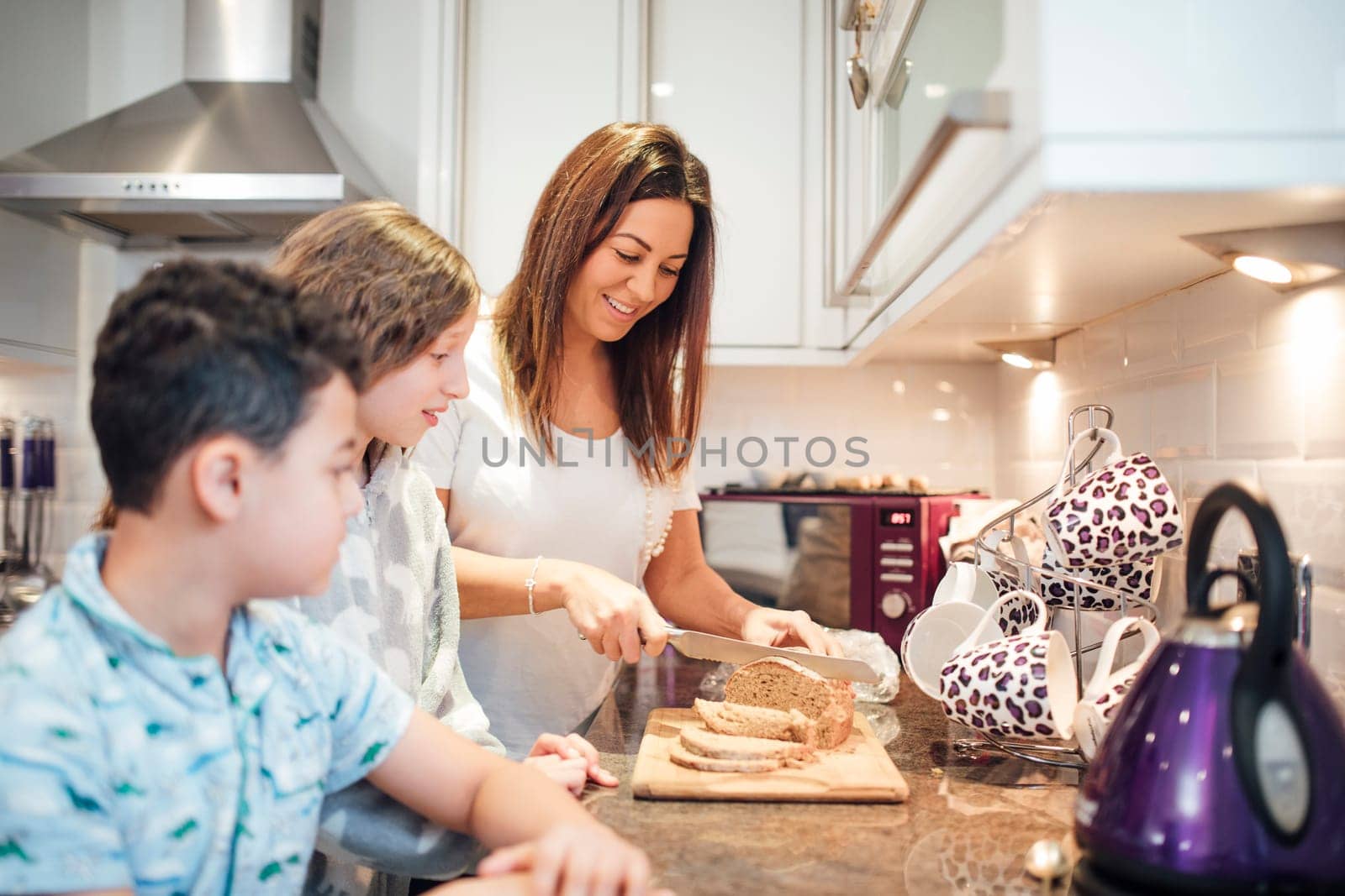 Mother is cutting slices of bread in the kitchen for her children to have toast for breakfast. They are watching eagerly.