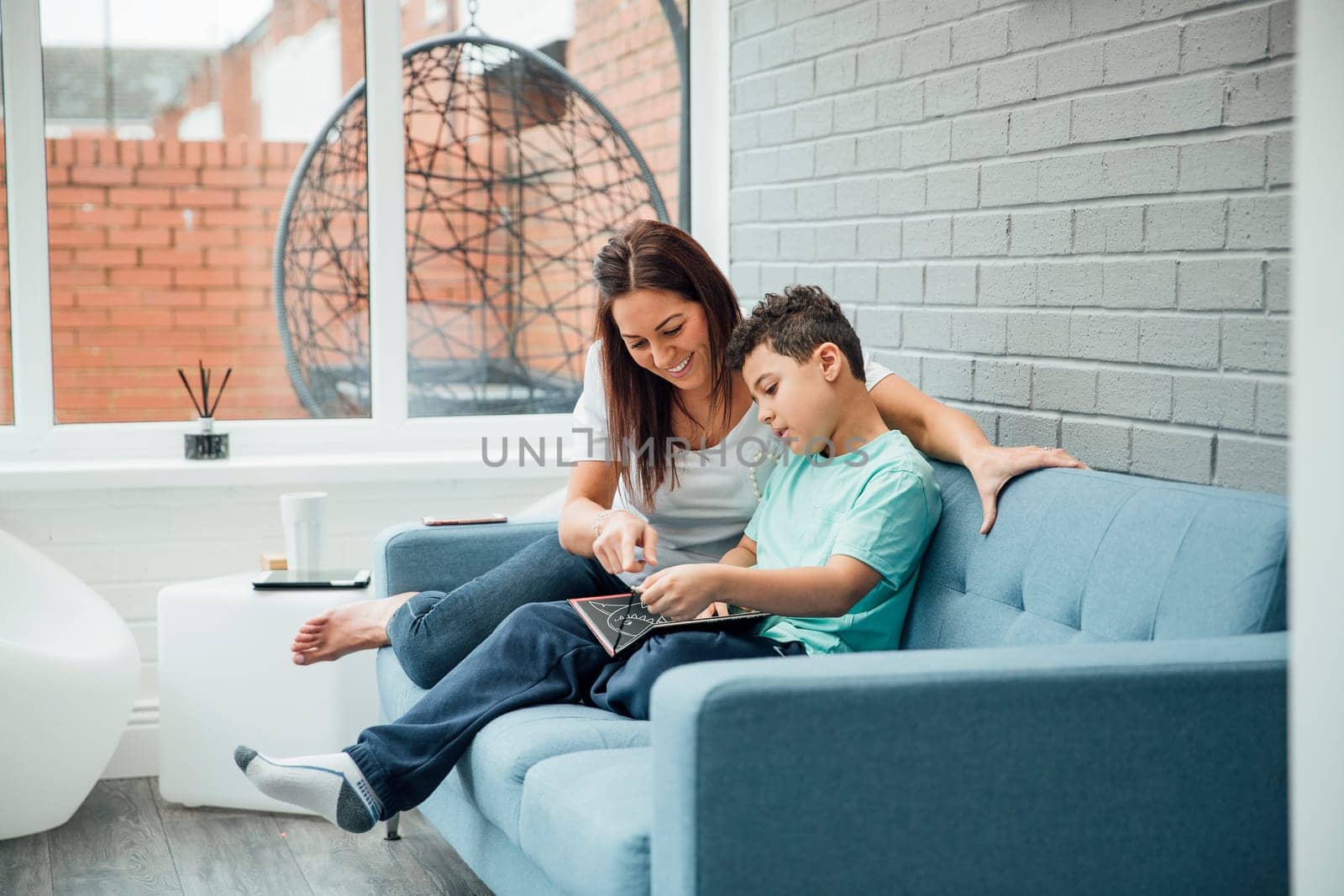 Little boy and his mother are reading a book together in the conservatory of their home.