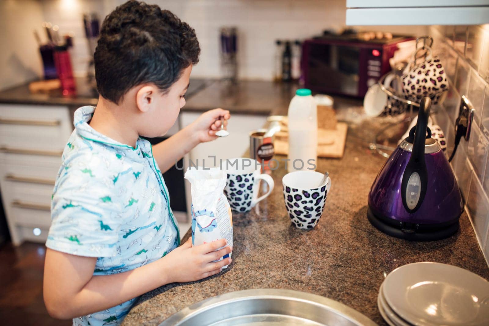 Little boy is helping to make tea for his parents at breakfast time. He is adding sugar to some cups.