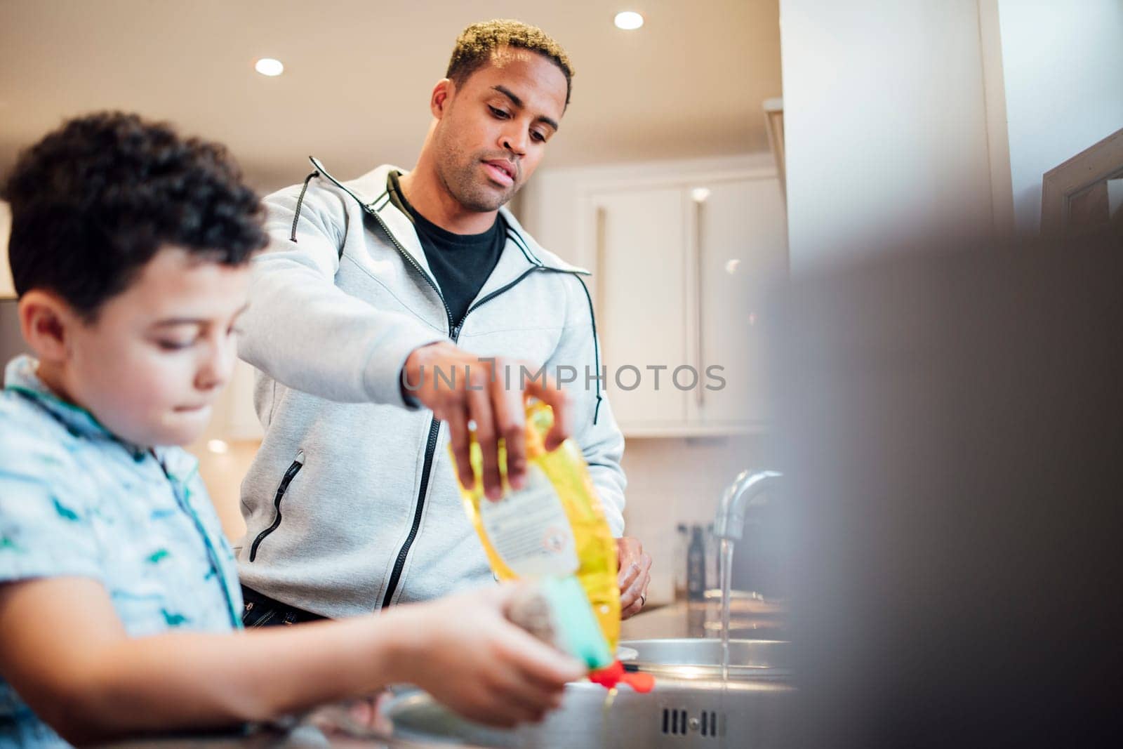 Little boy and his father are washing the dishes together at home. They are adding washing-up liquid to the sink.