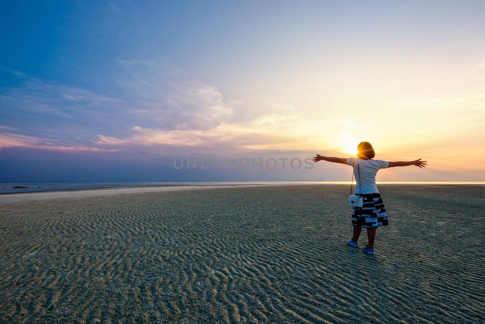 Happy woman tourist standing arms outstretched on the beach watching the beautiful natural landscape of colorful sky and sea during sunset at Nathon Sunset Viewpoint in Samui island, Surat Thani, Thailand
