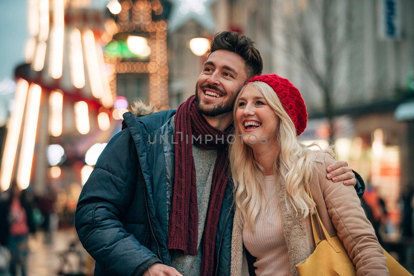 Front view of a man with his arm around his girlfriend outside. The couple are outside on a city street . There is a fair groung ride in the background.