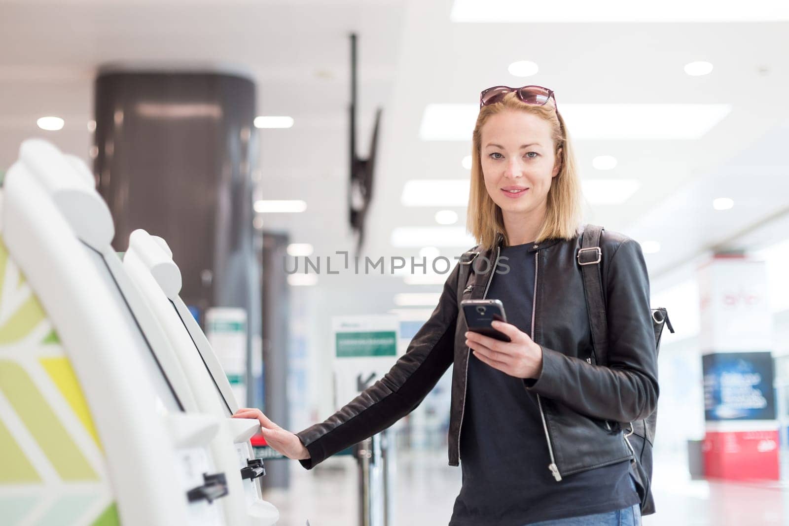 Casual caucasian woman using smart phone application and check-in machine at the airport getting the boarding pass. Model smiling, looking at camera. Modern technology on airport.
