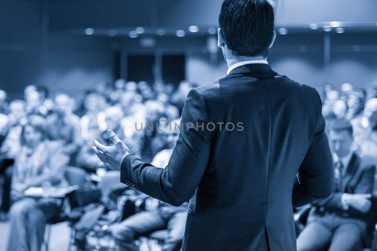 Speaker giving a talk on corporate business conference. Unrecognizable people in audience at conference hall. Business and Entrepreneurship event. Blue toned grayscale image.