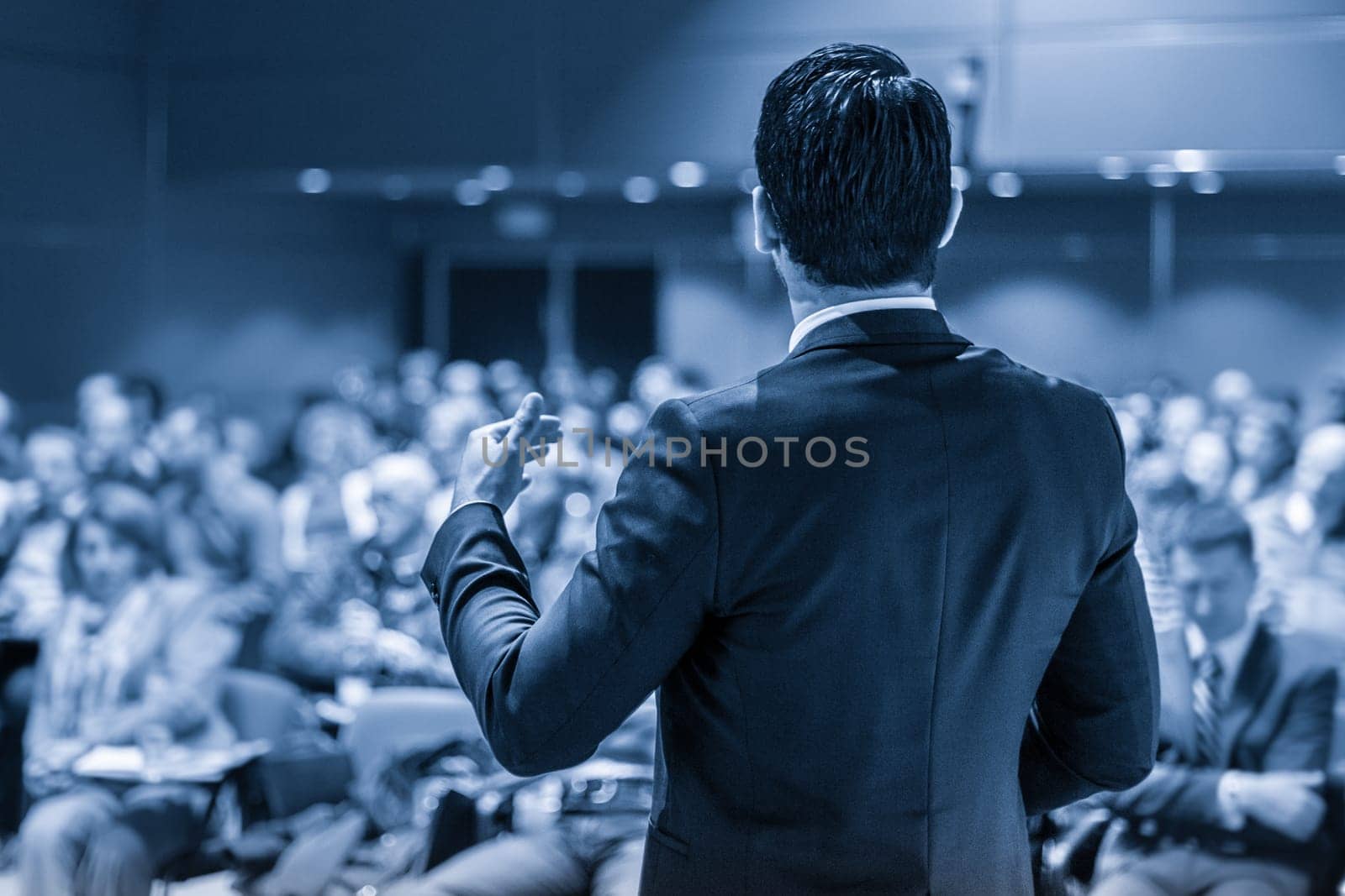 Speaker giving a talk on corporate business conference. Unrecognizable people in audience at conference hall. Business and Entrepreneurship event. Blue toned grayscale image.