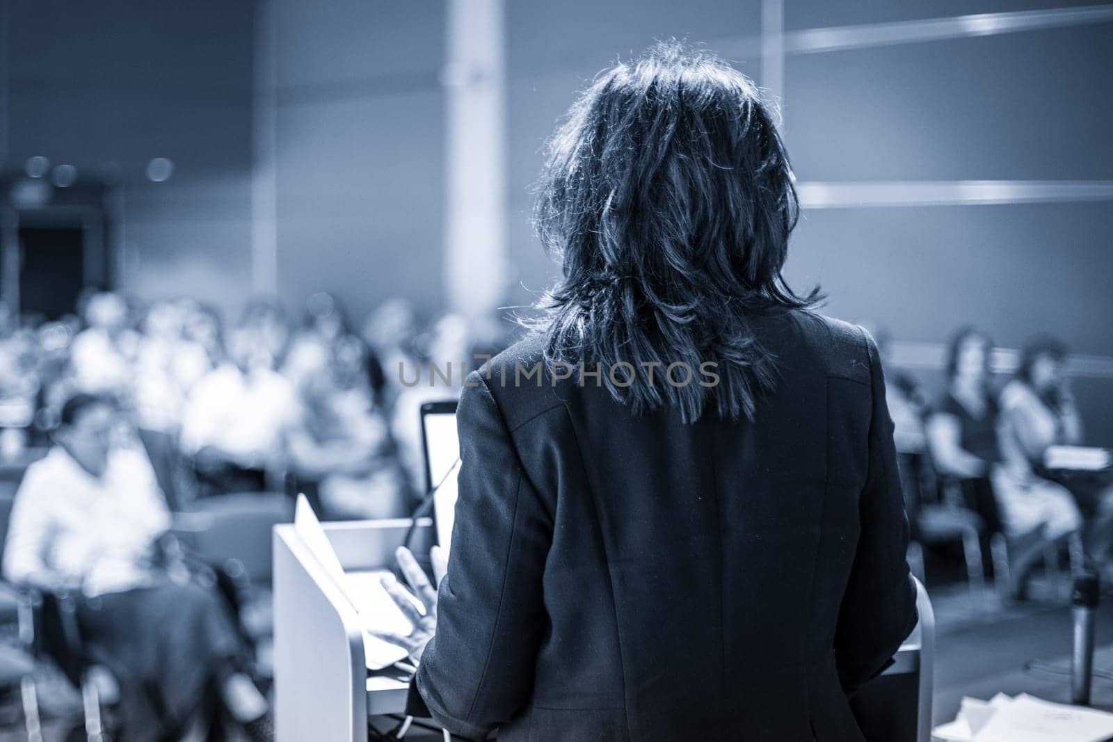 Female speaker giving a talk on corporate business conference. Unrecognizable people in audience at conference hall. Business and Entrepreneurship event. Black and white, blue toned image.