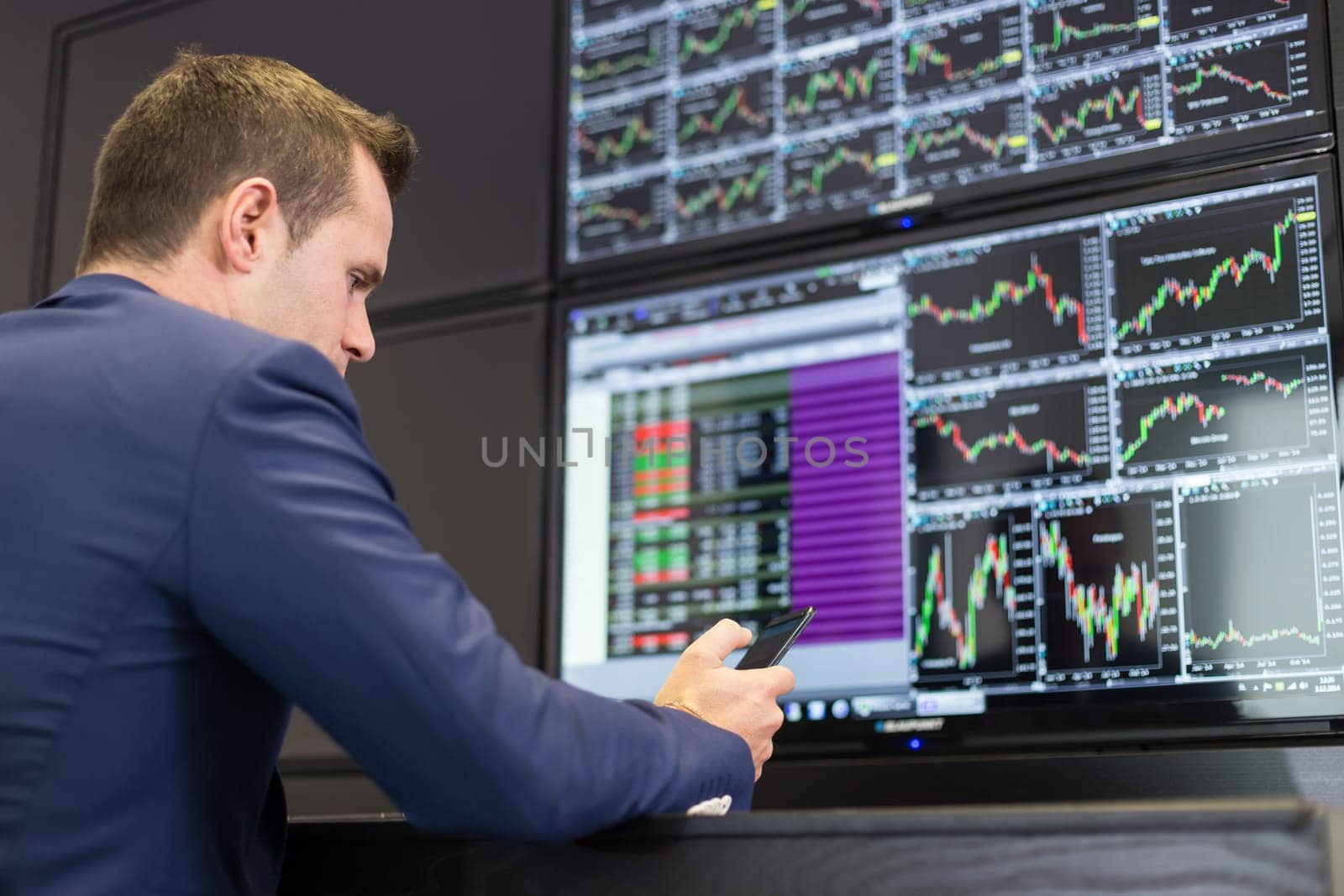 Businessman with cell phone trading stocks. Stock analyst looking at graphs, indexes and numbers on multiple computer screens. Stock trader evaluating economic data. Blue toned greyscale image.