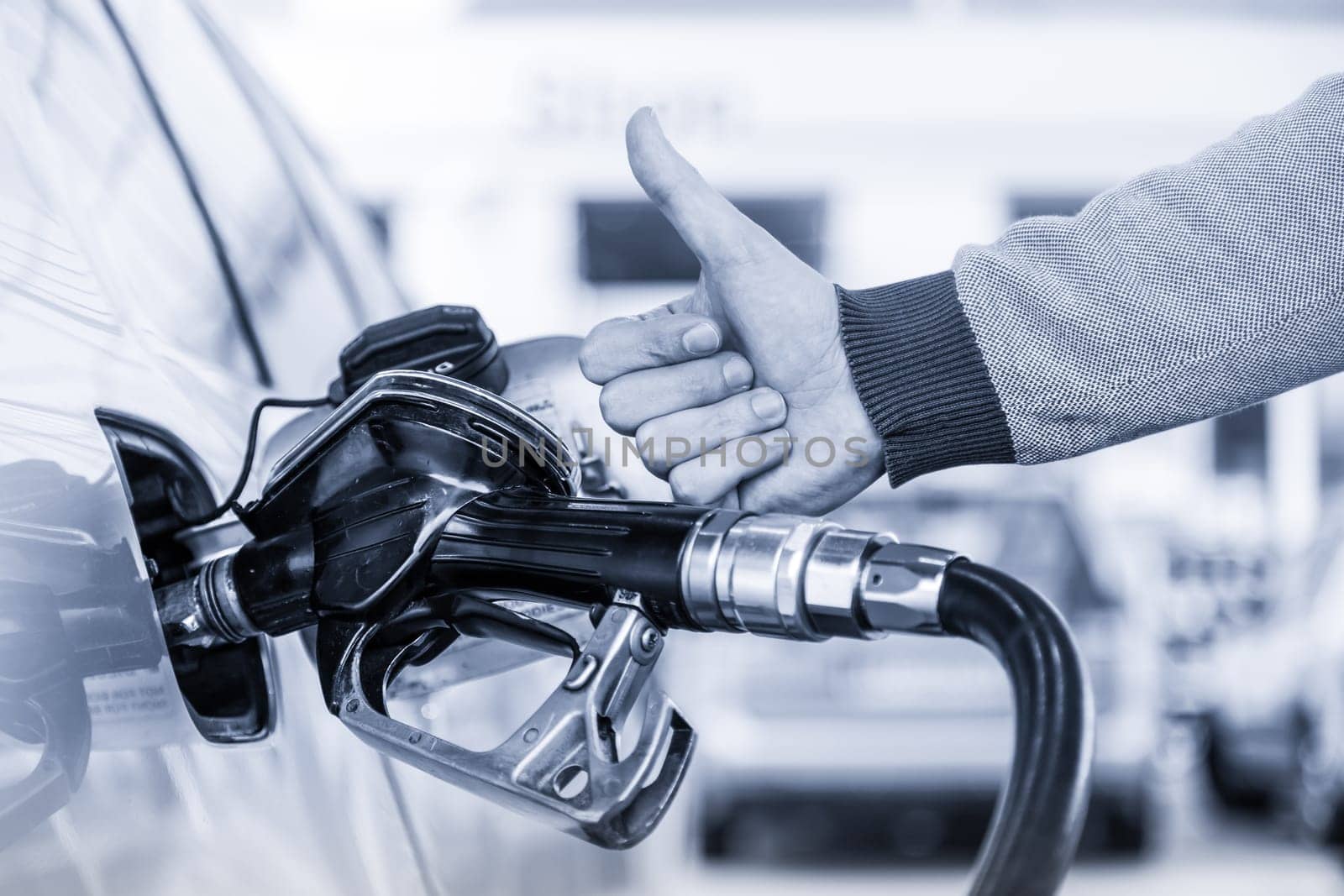 Petrol or gasoline being pumped into a motor vehicle car. Closeup of mans hand showing thumb up gesture, pumping gasoline fuel in car at gas station. Greyscale blue toned image.