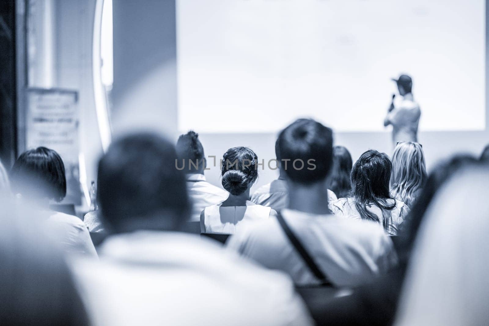 Speaker giving a talk in conference hall at business event. Focus on unrecognizable people in audience. Business and Entrepreneurship concept. Blue toned greyscale image.
