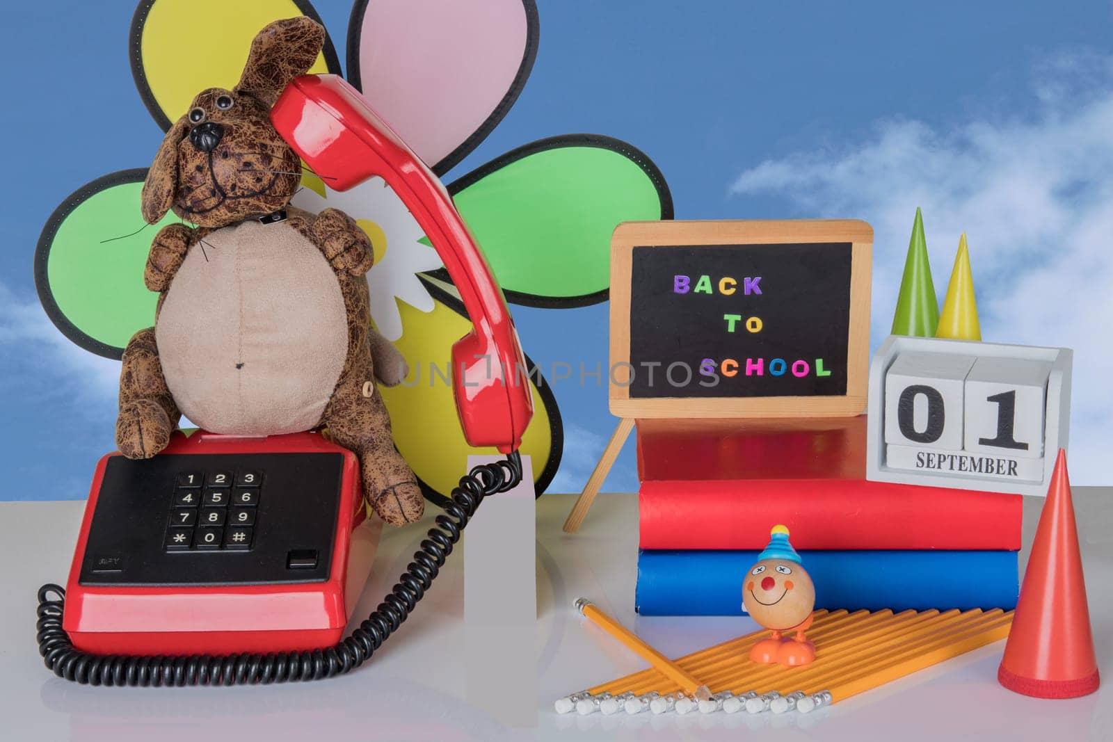 Colorful school equipment with funny dog and toy clown on white table against natural blue background. Back to school written with colorful letters on blackboard. A wooden calendar shows the 01 september if starts school. Back to school.