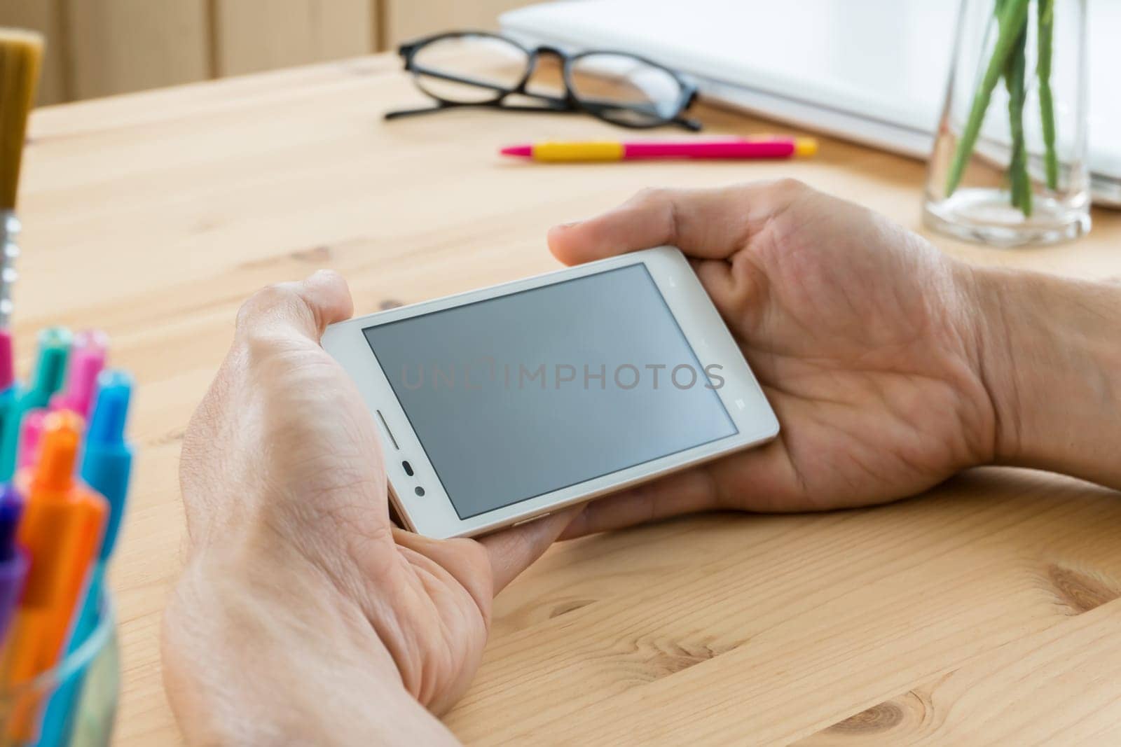 Smartphone or mobile phone on hand of human horizontal close up view. Smartphone or mobile phone and pen and eyeglasses and laptop or notebook on .wood table in home office. Business or technology concept for design.