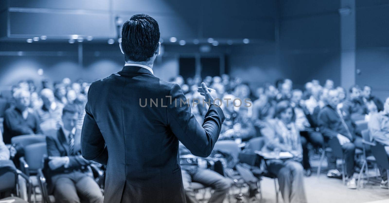 Speaker giving a talk on corporate business conference. Unrecognizable people in audience at conference hall. Business and Entrepreneurship event. Blue toned grayscale image.