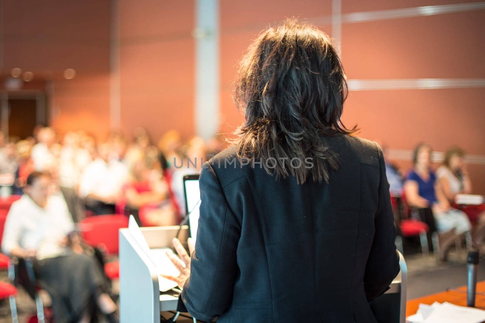 Female speaker giving a talk on corporate business conference. Unrecognizable people in audience at conference hall. Business and Entrepreneurship event.