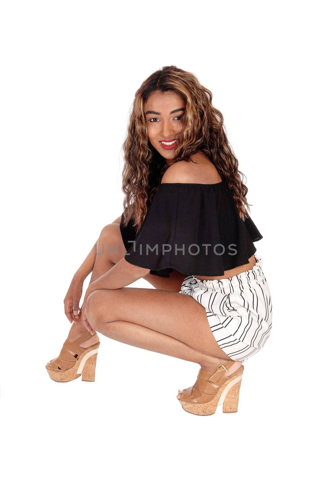 A happy lovely woman in a black blouse and white shorts crouching on.the floor whit her curly brunette hair, isolated foe white background.